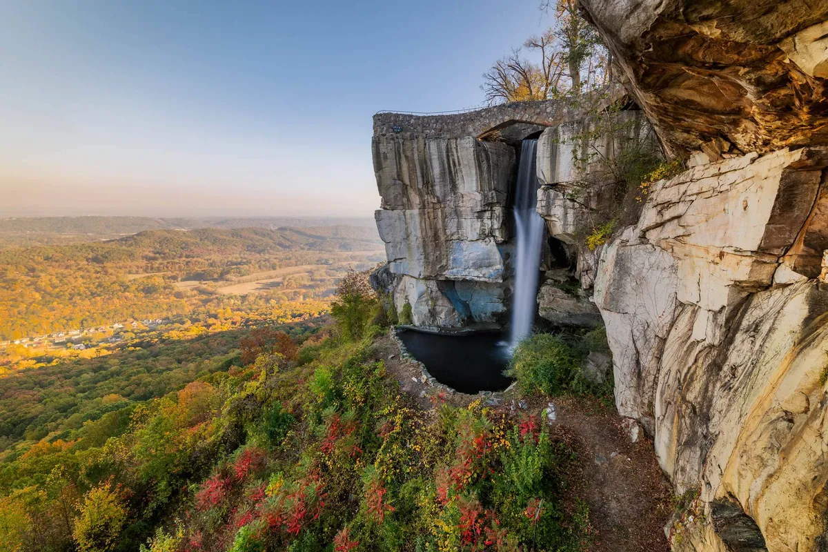 A picturesque waterfall pours over rock formations into a valley, framed by rich autumn foliage. The sweeping view hints at hiking trails, picnic spots, and the tranquility of nature.