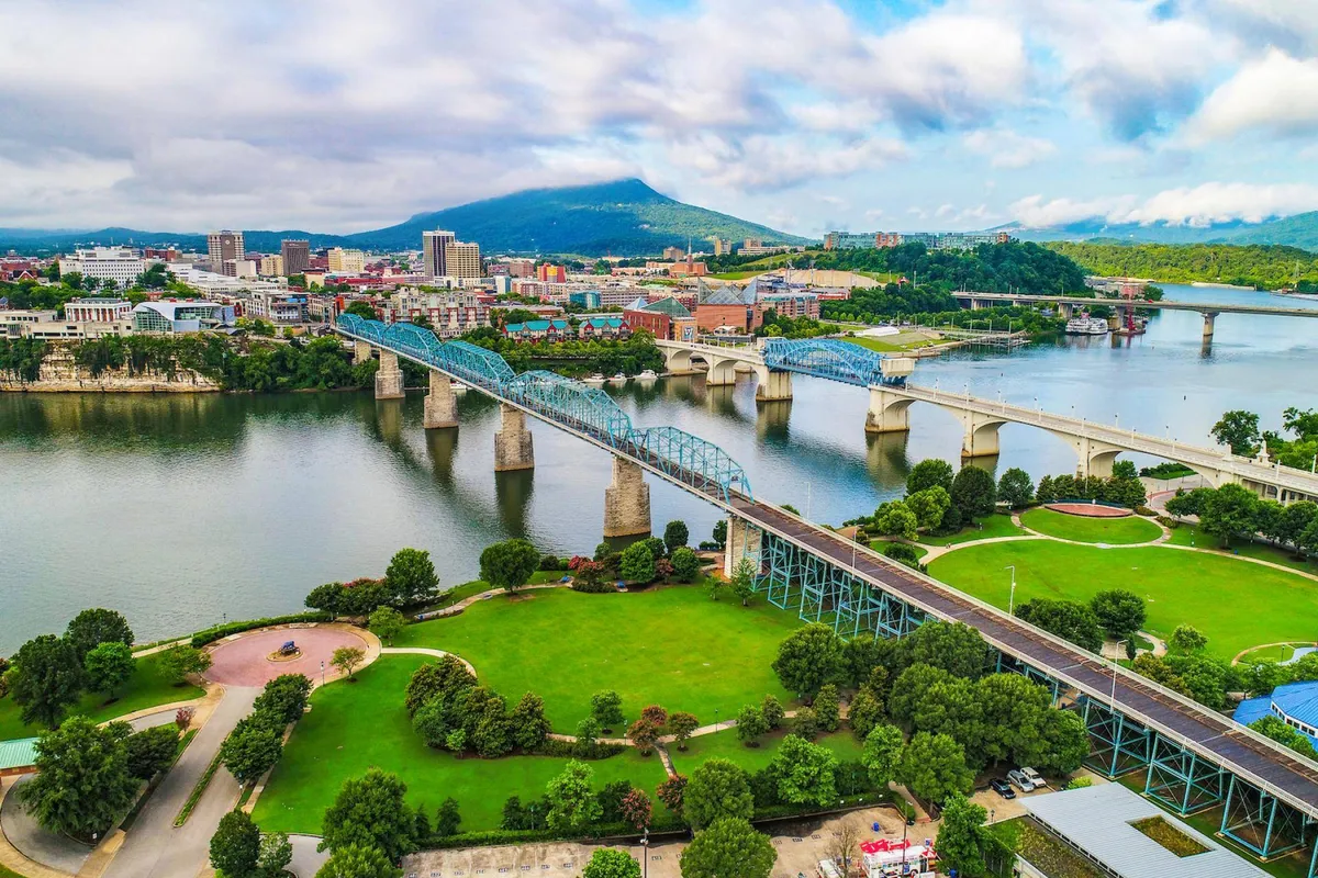 This image features a lively cityscape with bridges, a river, and a mountain backdrop, capturing the energetic vibe of Chattanooga. The scene is bustling with activities like walking and biking, and the inviting atmosphere is enhanced by sunset lighting and visible parks—ideal for community events and celebrations.