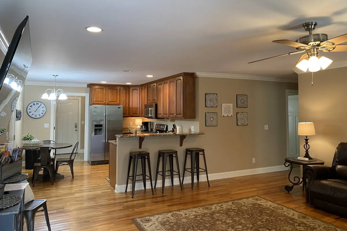 A cozy kitchen island with stools, adjacent to a dining area and living room. Decorative wall art adds warmth, and the setup encourages family and friends to gather around for casual snacks, conversations, or celebrations. The island is clearly stocked for easy meal prep and informal dining.