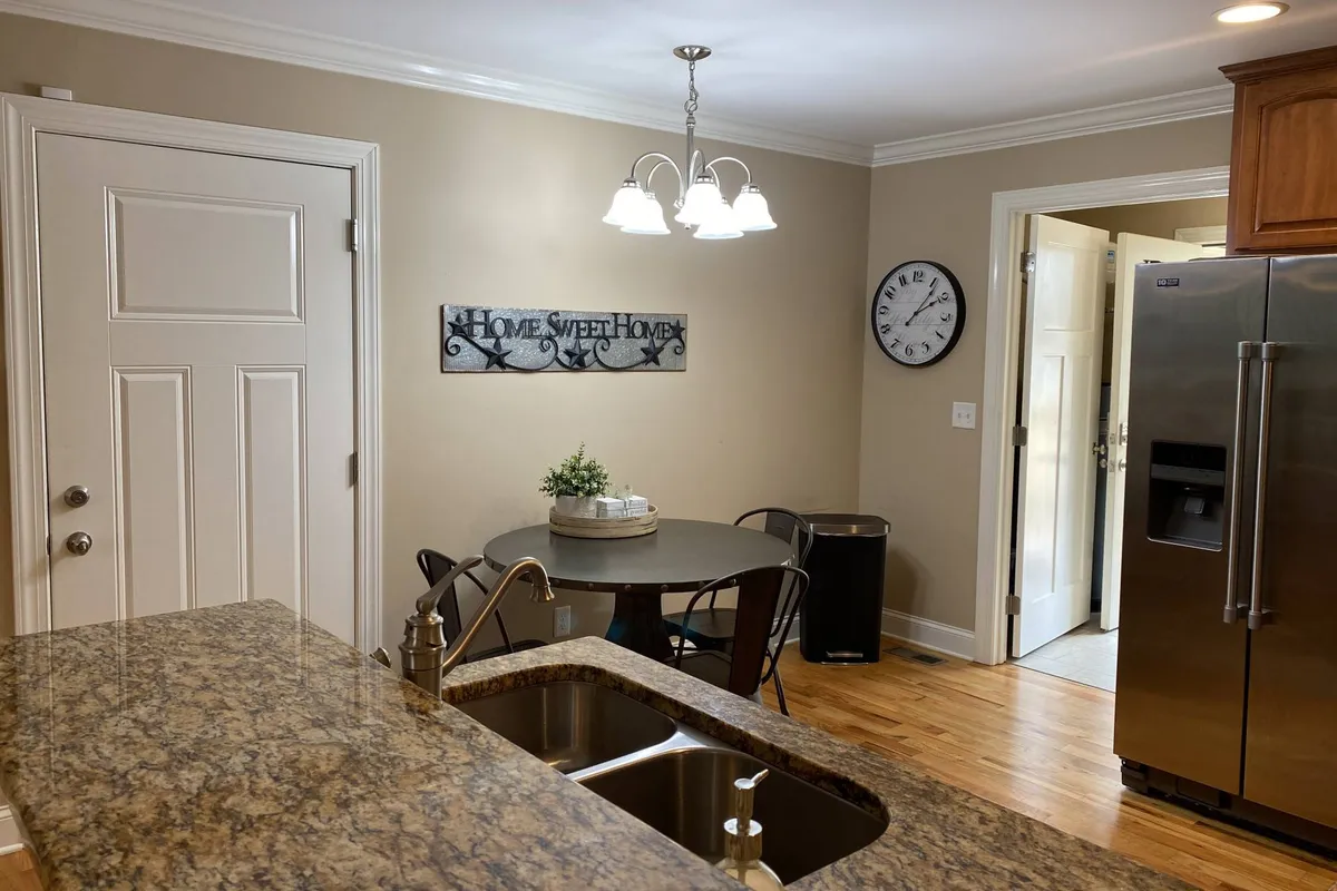 A view featuring a kitchen island with a sink, a round dining table, a 'Home Sweet Home' sign, and a wall clock. The space feels open and functional, suitable for both quiet moments and gatherings.